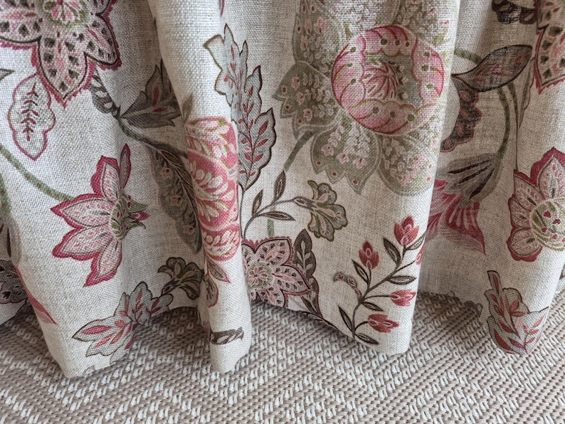 May include: Close-up of a floral fabric with a beige background. The fabric features large, stylized flowers in shades of pink, red, and olive green. The design includes detailed leaves and stems, creating a textured, botanical pattern. The fabric is draped, showing its folds and texture.