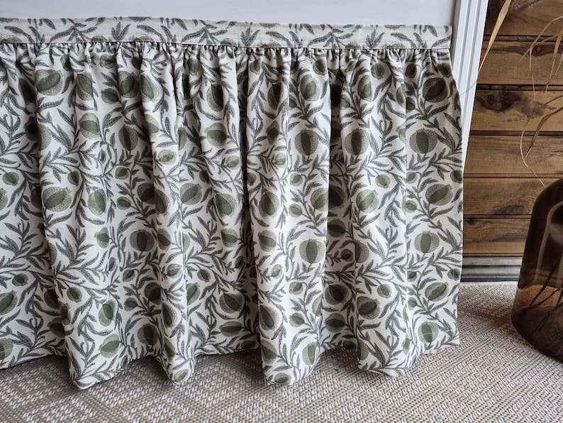 May include: A gathered valance with a botanical pattern. The fabric features a repeating design of green and grey leaves and fruit on a white background. The valance is hanging in front of a wooden wall.