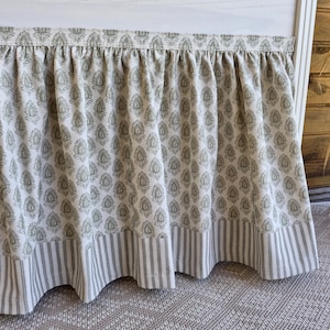 May include: A white bed skirt with a repeating olive green floral pattern. The bottom edge features a border of vertical gray and white stripes. The fabric is gathered at the top, creating a ruffled effect.