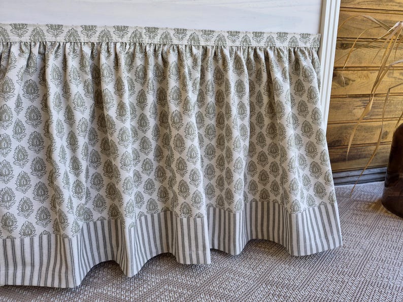May include: A white bed skirt with a repeating olive green floral pattern. The bottom edge features a border of vertical gray and white stripes. The fabric is gathered at the top, creating a ruffled effect.