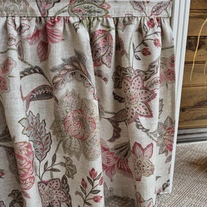May include: Close-up of a floral patterned fabric in shades of pink, brown, and green on a beige background. The fabric is gathered at the top, creating soft folds. The design features large, stylized flowers and leaves.