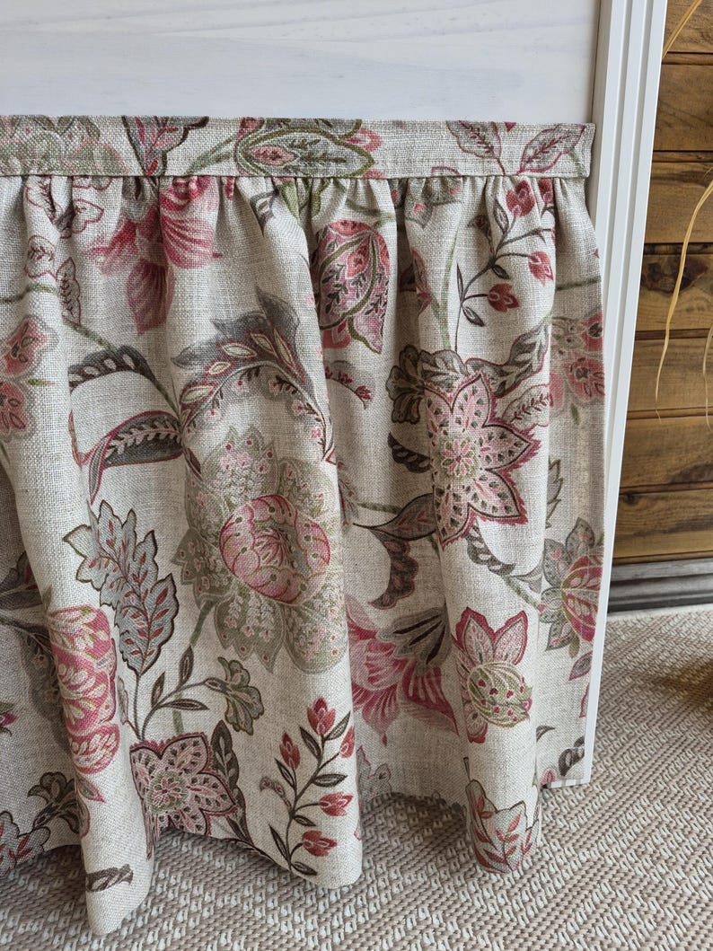 May include: Close-up of a floral patterned fabric in shades of pink, brown, and green on a beige background. The fabric is gathered at the top, creating soft folds. The design features large, stylized flowers and leaves.