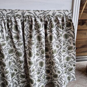 May include: A gathered fabric curtain featuring a repeating botanical pattern of sage green pomegranates and dark grey leafy branches on an off-white background. The curtain has a straight top header and hangs in soft vertical folds.