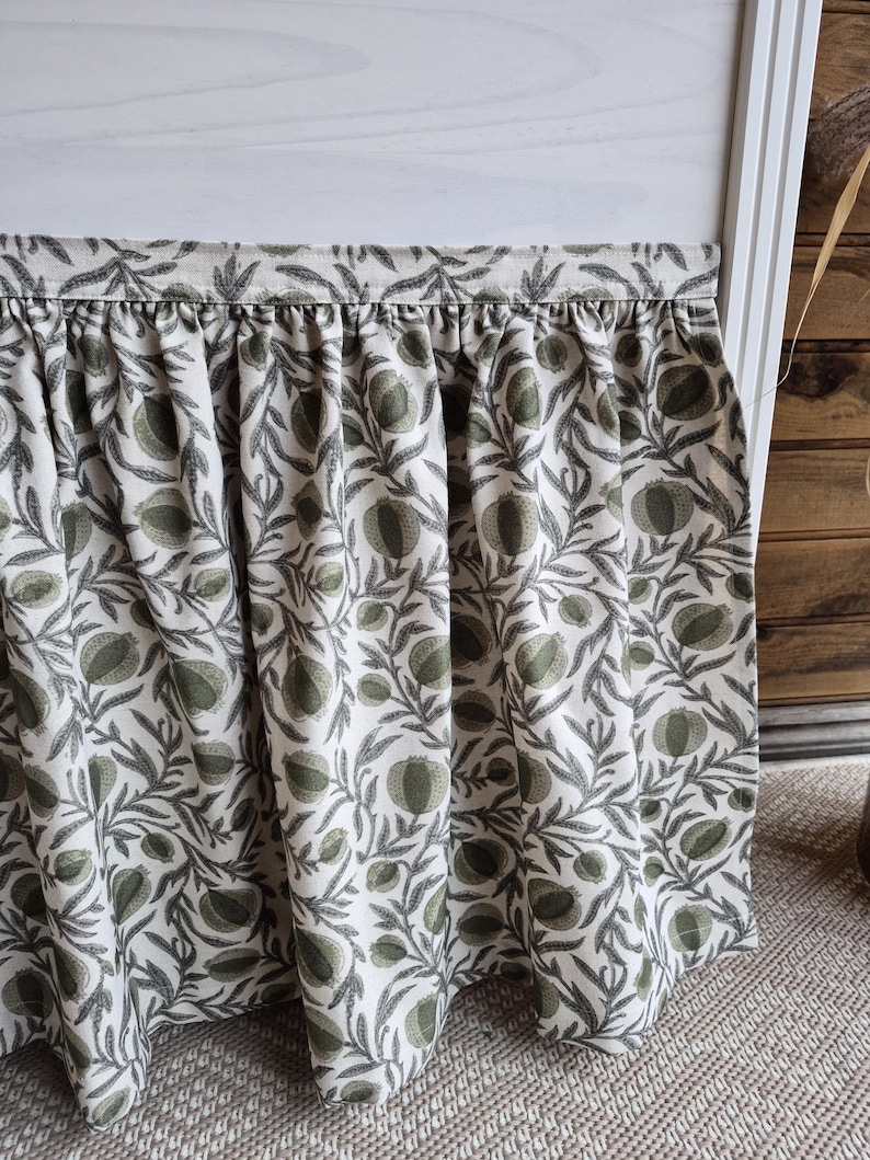 May include: A gathered fabric curtain featuring a repeating botanical pattern of sage green pomegranates and dark grey leafy branches on an off-white background. The curtain has a straight top header and hangs in soft vertical folds.