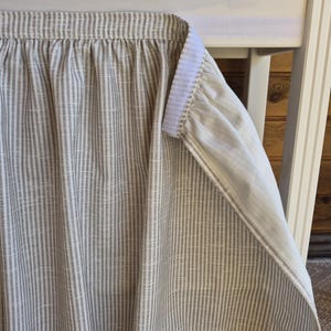 Könnte beinhalten: Ein beigefarbener und weiß gestreifter Stoffrock, möglicherweise für einen Tisch oder ein Bett, mit einem gerafften oberen Rand. Der Rock ist mit einem Klettverschluss an einer weißen Oberfläche befestigt. Der Stoff hat ein strukturiertes Aussehen.