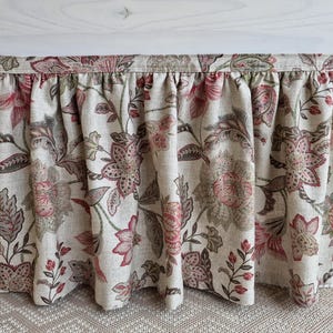 May include: A floral valance with a gathered design. The fabric features a pattern of large pink and gray flowers with green leaves on a beige background. The valance is hanging in front of a white structure.