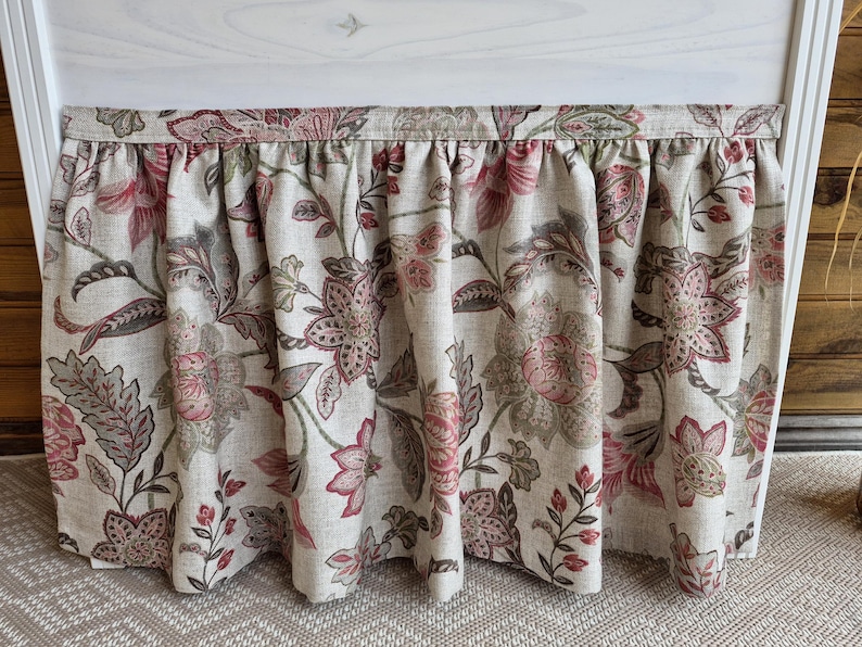 May include: A floral valance with a gathered design. The fabric features a pattern of large pink and gray flowers with green leaves on a beige background. The valance is hanging in front of a white structure.