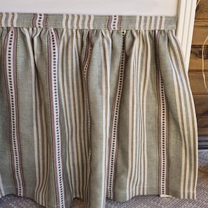 May include: A bed skirt featuring vertical stripes in green, white, brown, and purple. The fabric is gathered at the top, creating soft folds. The skirt is designed to hang from a bed frame.