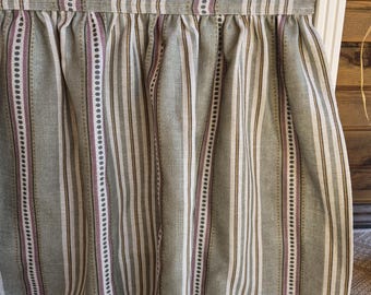May include: A bed skirt with vertical stripes in shades of green, white, brown, and purple. The fabric is gathered at the top, creating soft folds. The skirt is designed to hang from a bed frame.
