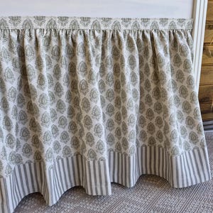 May include: A gathered bed skirt with a floral pattern in olive green on a cream background. The bottom edge features a striped border in the same colors. The fabric is draped over a white bed frame.