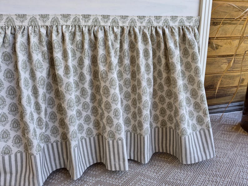 May include: A gathered bed skirt with a floral pattern in olive green on a cream background. The bottom edge features a striped border in the same colors. The fabric is draped over a white bed frame.
