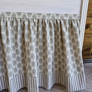 May include: A bed skirt with a cream background and a repeating olive green floral pattern. The bottom edge features a striped border in the same color scheme. The skirt is gathered at the top and hangs down, creating a decorative element for the bed.