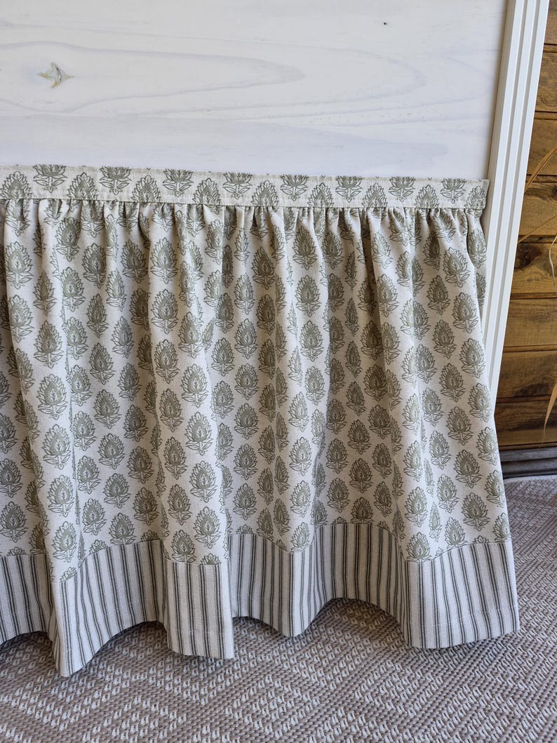 May include: A bed skirt with a cream background and a repeating olive green floral pattern. The bottom edge features a striped border in the same color scheme. The skirt is gathered at the top and hangs down, creating a decorative element for the bed.