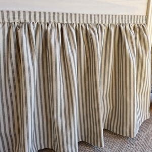 Puede incluir: Una falda de cama con diseño fruncido, con rayas verticales en crema y gris. La tela tiene una apariencia texturizada. Este textil de dormitorio añade una estética clásica y rústica.