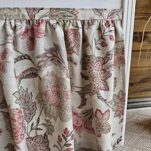 May include: A floral valance with a cream background and a pattern of pink, red, and gray flowers and leaves. The fabric is gathered at the top, creating soft folds. The valance is likely for a window treatment.