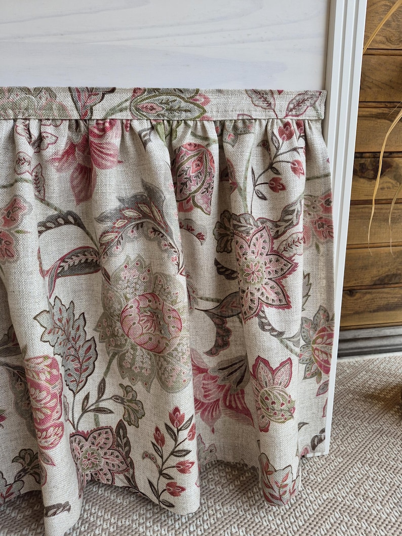 May include: A floral valance with a cream background and a pattern of pink, red, and gray flowers and leaves. The fabric is gathered at the top, creating soft folds. The valance is likely for a window treatment.