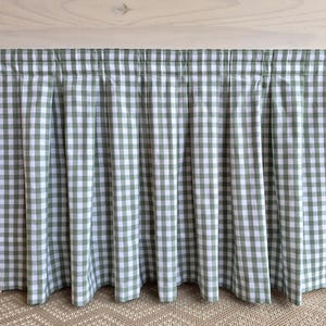 May include: A green and white gingham valance with a pleated design. The fabric is draped over a white wooden frame, creating a classic, country-style aesthetic. The valance adds a touch of charm to any window.