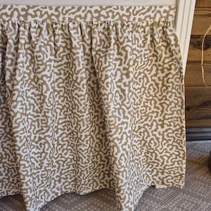 May include: A beige and white bed skirt with a repeating, organic pattern. The fabric is gathered at the top, creating soft folds. The pattern resembles a network of interconnected shapes, adding visual interest to the bedroom decor.