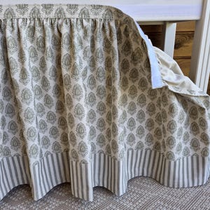 May include: A cream-colored crib skirt with a repeating floral pattern in muted green and a striped border. The skirt is attached with a white velcro strip and hangs in soft folds. The fabric has a textured appearance.