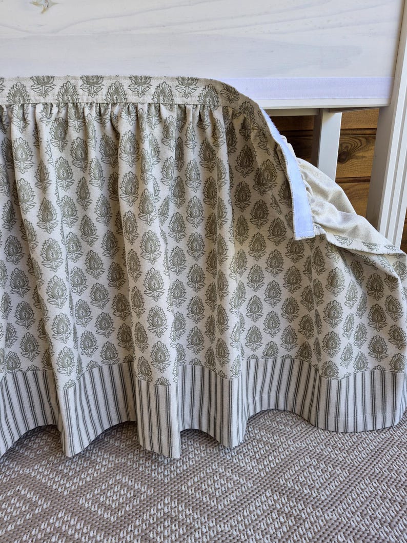 May include: A cream-colored crib skirt with a repeating floral pattern in muted green and a striped border. The skirt is attached with a white velcro strip and hangs in soft folds. The fabric has a textured appearance.
