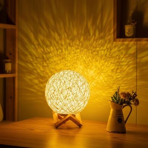 Puede incluir: Una lámpara esférica tejida con un brillo cálido y suave se encuentra sobre un soporte de madera. La lámpara proyecta un patrón de luz texturizado en la pared. A la derecha de la lámpara hay una pequeña jarra amarilla con flores.