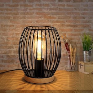 Puede incluir: Lámpara de mesa de jaula de metal negro con una bombilla cálida y brillante. La lámpara se asienta sobre una base de madera clara y un soporte de metal negro. La lámpara está sobre una mesa de madera con un fondo de pared de ladrillo. Luminaria decorativa.