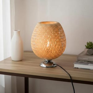 May include: A woven rattan table lamp with a chrome base, emitting a warm glow. The lamp has an egg-shaped shade with a diamond pattern. A white vase and a small succulent plant sit nearby on a wooden table.