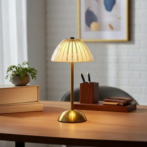 Könnte beinhalten: Eine Messing-Schreibtischlampe mit geripptem Glasschirm. Die Lampe hat einen abgerundeten Sockel und einen schlanken Stiel. Der Schirm ist cremefarben mit vertikalen Linien. Die Lampe steht auf einem Holzschreibtisch.