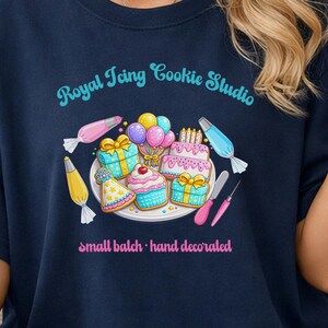 May include: Navy blue t-shirt featuring a colorful illustration of decorated cookies, gifts, and a cake. The text "Royal Icing Cookie Studio" is arched above the design, with "small batch • hand decorated" below.