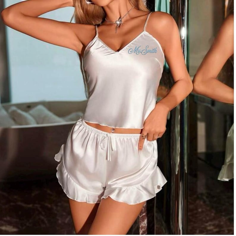May include: White satin pajama set. The set includes a camisole top with thin straps and a V-neck, and matching shorts with a ruffled hem. The top has the name "Ms. Smith" embroidered on the front. The shorts have a drawstring waist.