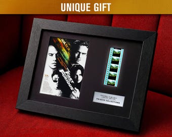 May include: Framed movie poster and film cell from "The Fast and the Furious." The black frame holds the movie poster, a strip of film, and a plaque. The poster features actors from the film. The film cell shows a series of images. The text "UNIQUE GIFT" is at the top.