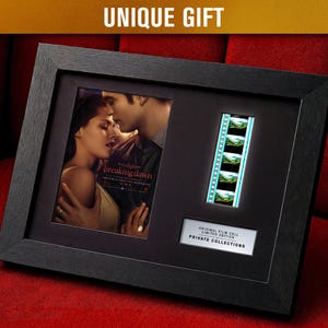 Pode incluir: Exibição emoldurada em preto com um pôster de filme e uma tira de filme. O pôster mostra um casal se abraçando, com o texto "Twilight Breaking Dawn". Uma tira de célula de filme é visível. A moldura inclui o texto "UNIQUE GIFT."