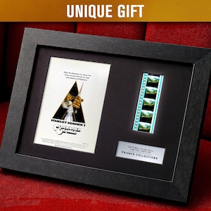 May include: Framed artwork featuring a movie poster and film strip from Stanley Kubrick's "A Clockwork Orange." The black frame holds the poster with a man holding a weapon, and a film strip with green and blue images. The text "UNIQUE GIFT" is at the top.