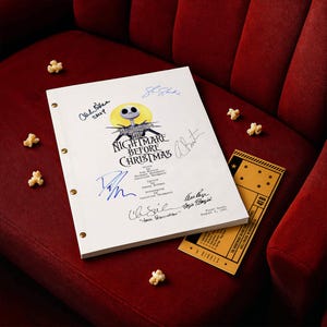 May include: A white script for "The Nightmare Before Christmas" with signatures, a movie ticket, and popcorn on a red velvet surface. The script features the film's title and a Jack Skellington illustration. The ticket is gold with black text.