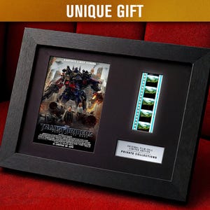 May include: Framed Transformers movie memorabilia. The black frame holds a movie poster and a strip of film cells. The poster features a battle scene with robots. The text "UNIQUE GIFT" is at the top. The film cells are illuminated.