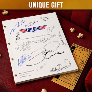 Peut inclure: Un scénario de film Top Gun blanc avec plusieurs signatures, le logo du film et le texte "UNIQUE GIFT". Un ticket de cinéma vintage et du pop-corn sont sur une surface en velours rouge.