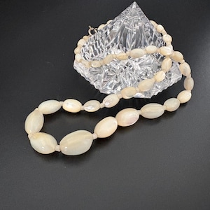 May include: A necklace composed of smooth, oval-shaped off-white beads with a pearlescent luster. The beads are strung together in a single strand, featuring a subtle variation in size, with larger beads positioned toward the front of the necklace.