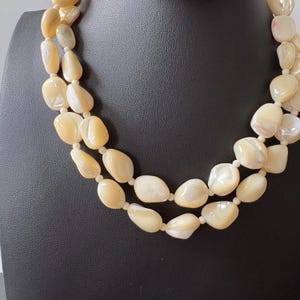 May include: A two-strand necklace featuring large, irregular, cream-colored beads with iridescent highlights. The beads are strung together, creating a layered effect. The necklace is displayed on a black mannequin.