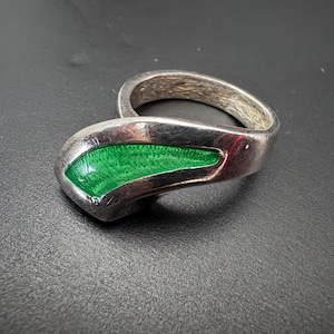 May include: A silver ring featuring a smooth, curved band with a prominent, elongated triangular inlay of vibrant green enamel. The metal surface shows a textured, brushed finish, creating a contrast with the glossy, bright green accent.