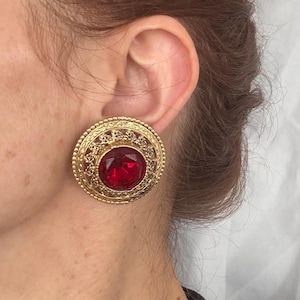 May include: A gold-toned, round clip-on earring with a large, faceted, red gemstone center. The earring features an ornate, filigree design around the gemstone and outer edge. The earring is clipped to an ear.