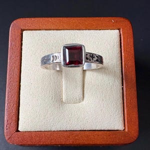 May include: A silver ring with a square, deep red gemstone set in the center. The band features etched designs on either side of the gemstone. The ring is displayed in a wooden jewelry box with a beige interior.