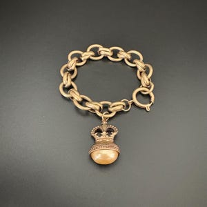 May include: A gold-toned chain link bracelet featuring a crown-shaped charm. The charm is adorned with a smooth, round, peach-colored bead at its base. The bracelet includes a circular spring ring clasp for secure fastening and closure.