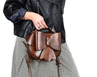 May include: A brown leather handbag with a large bow detail. The bag has a long strap and is worn over the shoulder.