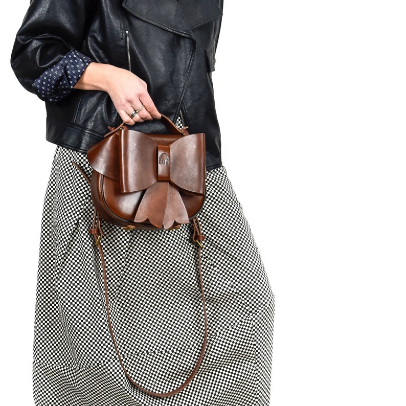 May include: A brown leather handbag with a large bow detail. The bag has a long strap and is worn over the shoulder.