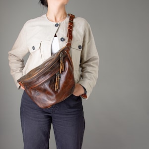 Könnte beinhalten: Eine braune Leder-Bauchtasche mit einem langen Riemen. Die Tasche hat einen Reißverschluss und eine Klappe mit einer Schnalle. Die Tasche wird von einer Person getragen, die eine hellbeige Jacke und eine blaue Jeans trägt.