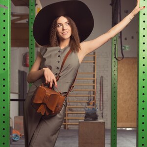 May include: A woman wearing a wide-brimmed black hat and a grey jumpsuit with a brown leather bag featuring a bow detail. She is standing in a gym setting with green metal bars in the background.