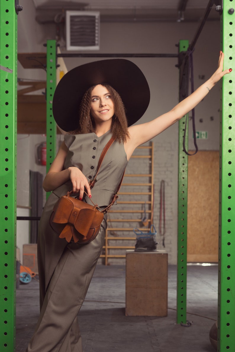 May include: A woman wearing a wide-brimmed black hat and a grey jumpsuit with a brown leather bag featuring a bow detail. She is standing in a gym setting with green metal bars in the background.
