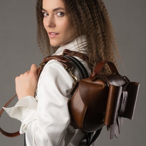 May include: A woman wearing a white shirt and black and white striped trousers is wearing a brown leather backpack with a unique bow detail. The backpack has a strap that goes over the shoulder and a handle on the top.