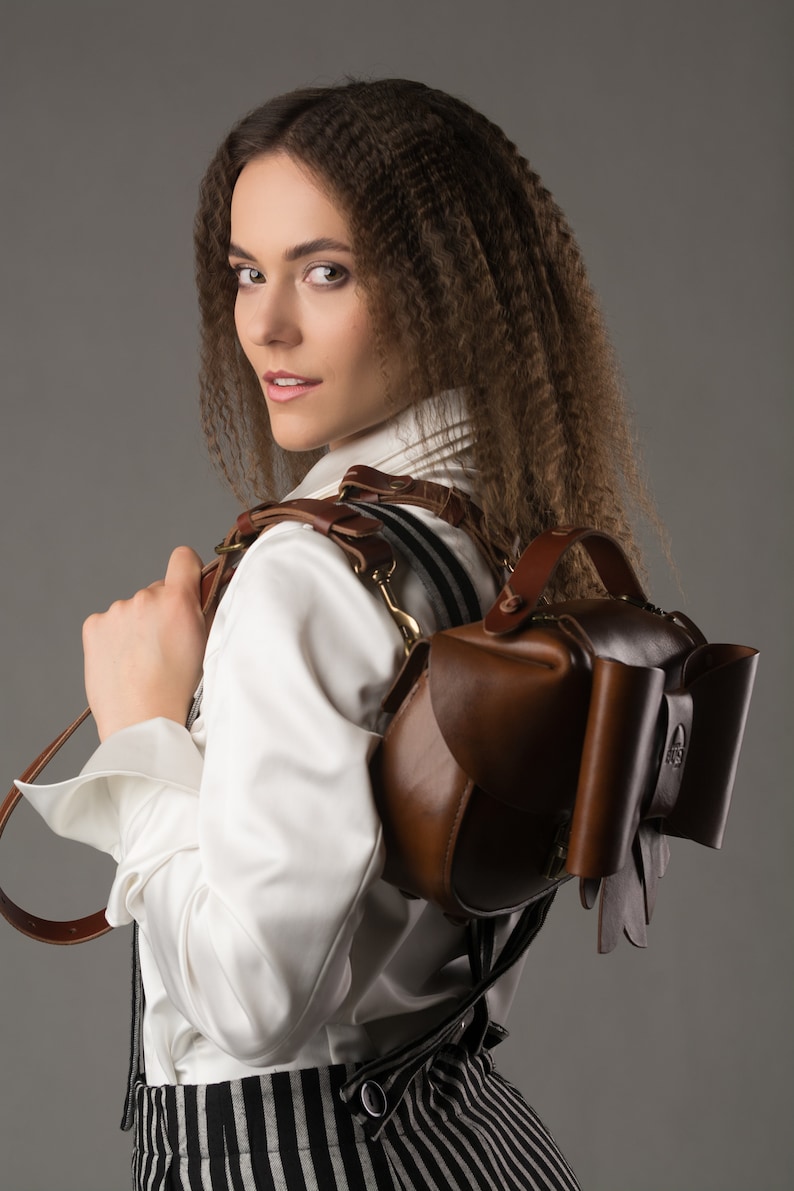 May include: A woman wearing a white shirt and black and white striped trousers is wearing a brown leather backpack with a unique bow detail. The backpack has a strap that goes over the shoulder and a handle on the top.