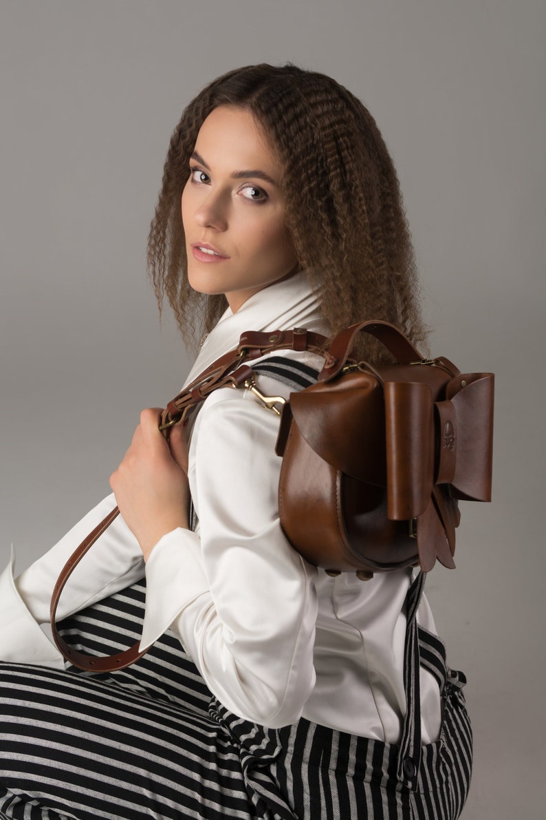 May include: A brown leather satchel with a large bow detail. The bag has a long strap and is worn over the shoulder.